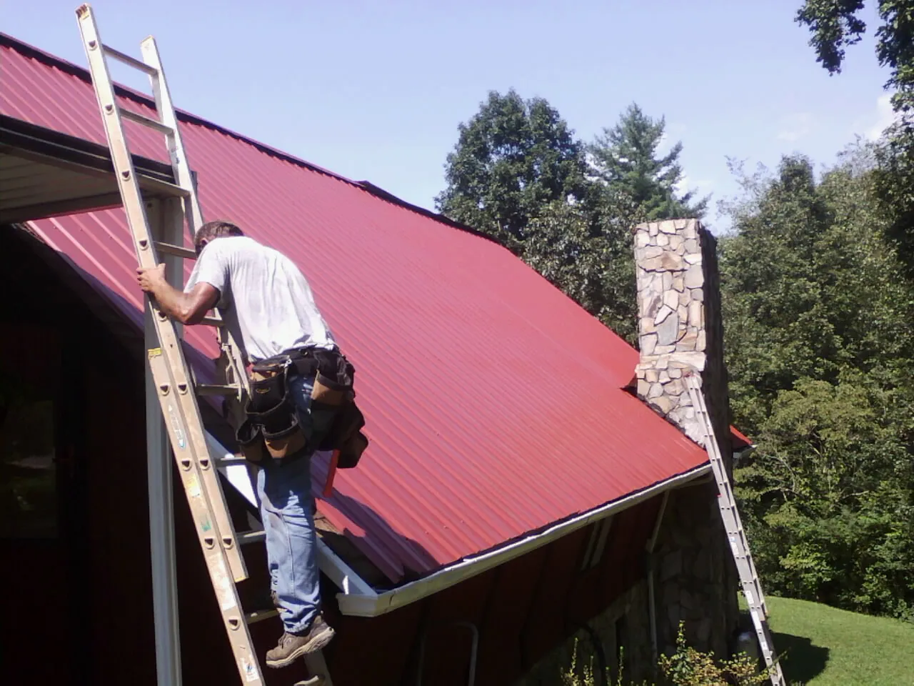 Roofer on a ladder beside a red metal roof and stone chimney — Soffit & Fascia Repair by Roofing Brothers in Yeagertown
